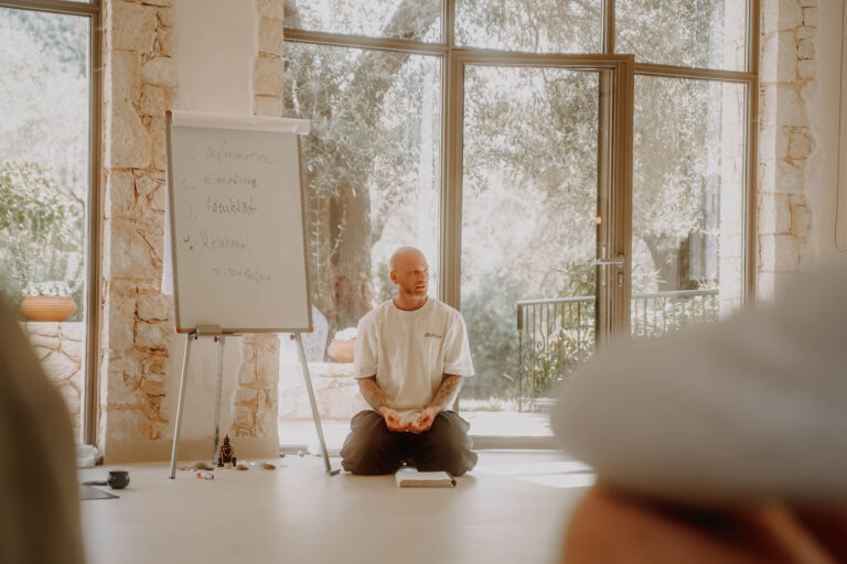 Theorie-Einheit in einem hellen Seminarraum: Ein Mann kniet vor einem Flipchart und spricht zur Gruppe, hinter ihm Fenster mit Blick auf Olivenbäume.