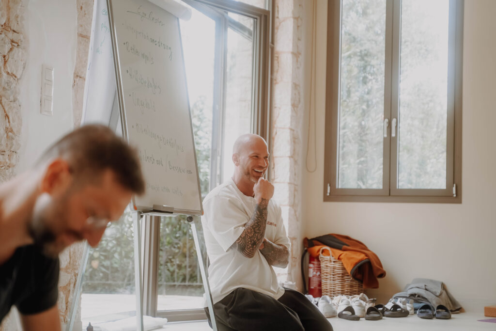 Seminarraum mit Flipchart: Ein Mann sitzt lachend vor dem Fenster, im Vordergrund unscharf ein Teilnehmer.